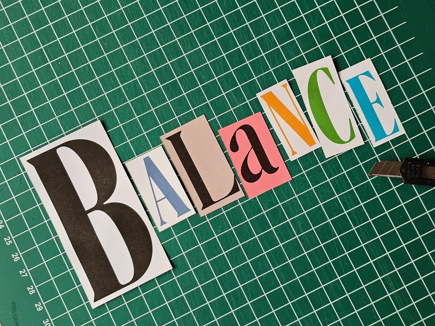 Multi-coloured cut-out letters spelling BALANCE arranged on a green cutting mat